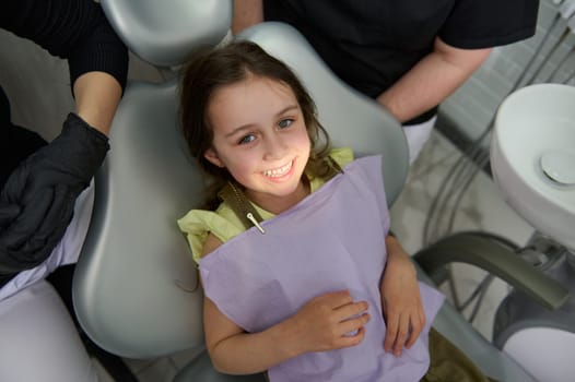 Adorable little child girl 5 years old, smiling a toothy smile looking at camera, sitting at dentist chair while visiting dental clinic for regular check-up. Pediatric dentistry. Dental oral and care