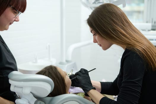 Female dentist hygienist using dental mirror and probe, checking child teeth during regular check-up in dentistry clinic. Little girl visiting doctor dentist hygienist. Oral care and hygiene concept