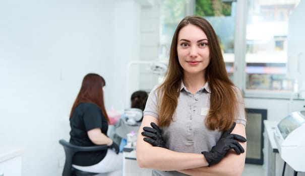 Confident portrait of a young female doctor dentist hygienist, healthcare worker, dental assistant posing with her arms folded, looking at camera, standing in her workplace in modern dentistry clinic
