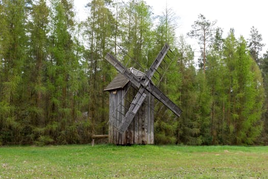 Old Wooden Mill In Meadow, Open Air. Blades Of Old Wooden Windmill In Rural Eastern Europe Area, Countryside. Green Forest On Background. Horizontal Landscape Plane High Quality Photo.