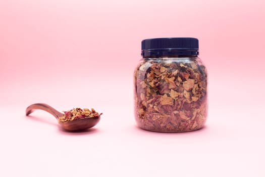 Jar, plastic container with dried rose petals, leaves. Wooden spoon filled with desiccated roses on pink background. Aromatic herbal beverage made from fragrant buds of flowers. Horizontal. Copyspace.