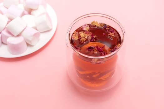 Healthy herbal rose petals, buds tea in double walled glass mug, cup. Pink white marshmallow plate. Pink background. Desiccated colorful flower petals herbal drink, beverage. Horizontal, top view.