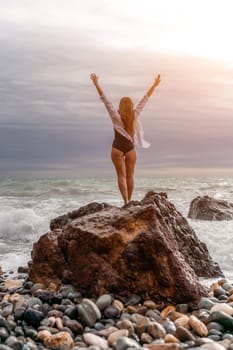 A beautiful girl in a white shirt and black swimsuit stands on a rock, big waves with white foam. A cloudy stormy day at sea, with clouds and big waves hitting the rocks