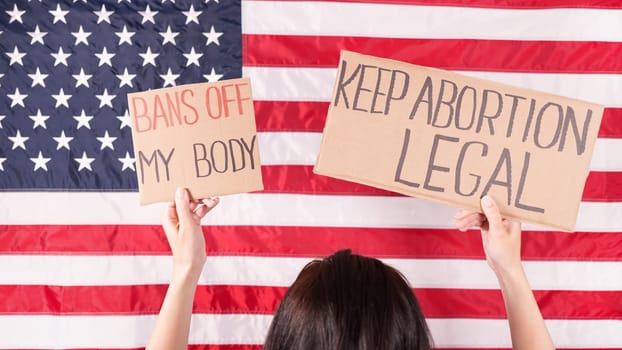 Young woman protester holds cardboard with Keep Abortion Legal and Bans Of My Body signs against USA flag on background. Girl protesting against anti-abortion laws. Womens rights freedom