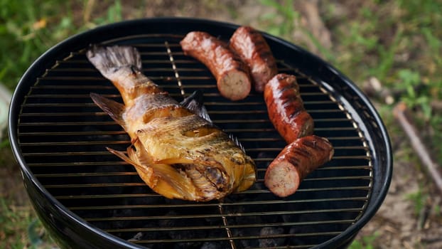 Top view of fresh grilled whole fish, skewer, sausages on round black charcoal grill, green grass background. Barbecue, grill and food concept. Camping with family