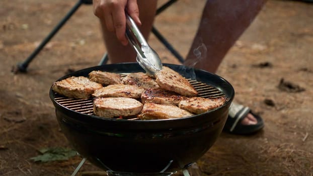 Unrecognized man is cooking BBQ stakes on charcoal grill in the forest. Camping food preparation. Fresh juicy stakes grilling on the barbeque