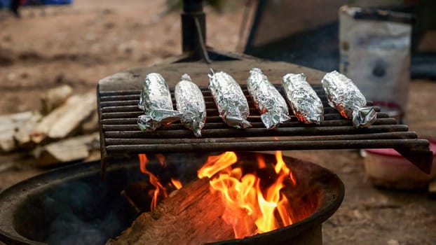 Close up shot of grilled barbecued sweetcorn cobs covered in foil in forest camping. Home cooking baked sweet corn. Summer picnic, concept of resting in fresh air. BBQ in garden