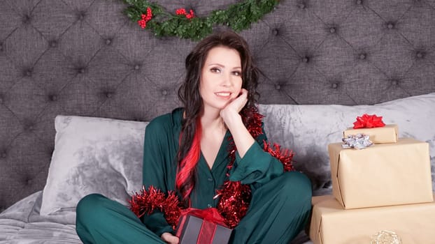 Happy young woman holding gift box for Christmas sitting on bed with xmas decorations, presents. Young girl wearing satin green pajama with holiday decorations at cozy living room