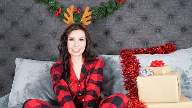 Happy young woman wearing red flannel pajama with holiday deer antlers sitting on bed with Christmas decorations, presents, copy space. Space for text