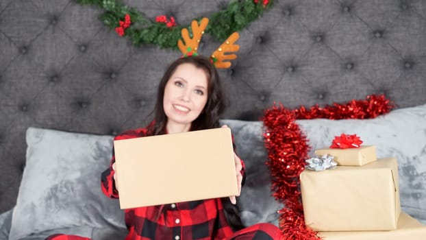 Happy young woman wearing red flannel pajama with holiday deer antlers and holding gift box with space for text sitting on bed with Christmas decorations, presents, copy space. Selective focus
