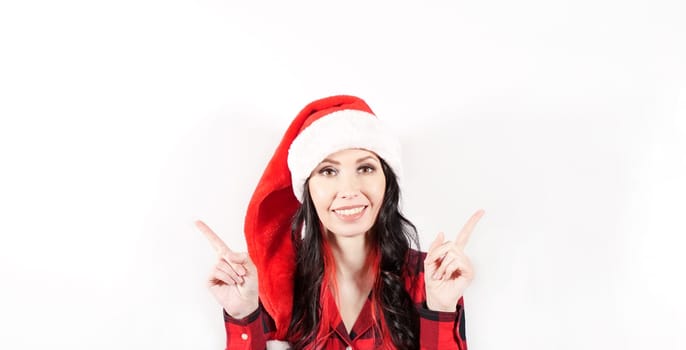 Smiling woman in red Christmas hat holding finger at empty Space for text for promo discount. Beautiful lady, Long curly hair, red santa hat, red pajama. Woman portrait isolated on white background
