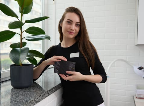 Charming young woman, doctor, aesthetician, beautician holding a black empty certificate in hands, looking at camera, standing by marble windowsill in a modern clinic, dressed in black medical uniform