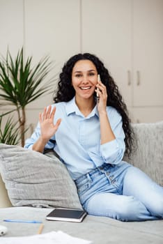 Beautiful young arab woman talking on the phone and waving hand in greeting. Girl sits on a gray sofa in a homely atmosphere in the workspace