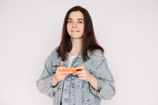 Portrait of a young woman in a casual denim jacket with a smartphone playing a game, isolated against a gray background, conveying indecision and insecurity.
