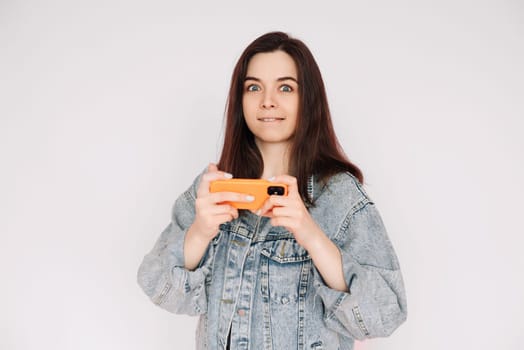 Indecisive and Insecure: Young Woman in Denim Jacket Playing Smartphone Game, Isolated on Gray Background. Emphasizing Uncertainty and Doubt.
