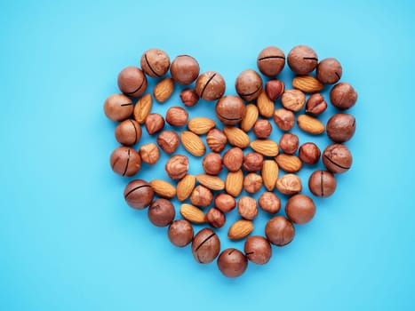 The concept of health care, cardio, proper nutrition, healthy fats. Various organic nuts forming heart shape on blue background. Tasty macadamia, hazelnut and almond. Top view and copy space.