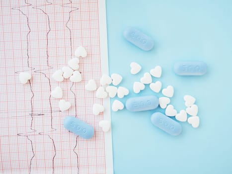 Healthcare concept. White medical cardiological pills in the form of heart and blue tablets on electrocardiogram and blue background. The drug for restoring health. Medical blog. Top view