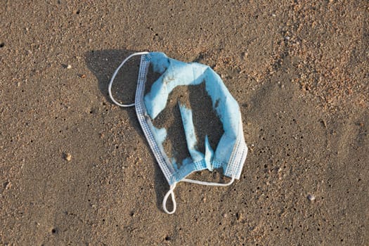 Discarded to beach of sea pale blue single-use face mask. Environmental and coast pollution. Used surgical mask thrown on the seashore threatening the health of ocean. Waste during COVID-19