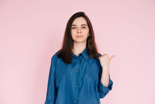 Young Businesswoman in Blue Shirt Pointing at Mockup, Symbolizing Office Promotion and New Opportunities: Professional Portrait on Pink Background.