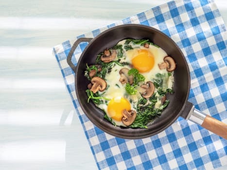 Idea for breakfast. Cooking pan with spinach, mushrooms, cheese and fried eggs. Healthy homemade dish for low carb diet on a light blue wooden table with linen towel. Close up, flat lay. Top view.