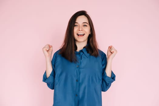 Euphoric Woman Celebrating Success, Raising Fists in Triumph: Isolated Pink Background Portrait. Joyful Woman Celebrating Achievement, Raising Fists in Excitement: Isolated Pink Background Portrait.