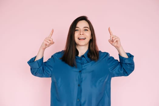 Vibrant Portrait of Energetic Young Woman Expressing Amazement with Open Mouth and Pointing Fingers Upward, Isolated on Pink Background.