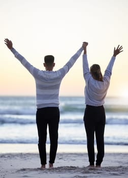 Couple, beach and hands up outdoor while happy at sunset for love, freedom and peace with calm ocean. Young man and woman together on vacation holiday at sea to relax, travel and connect in nature.