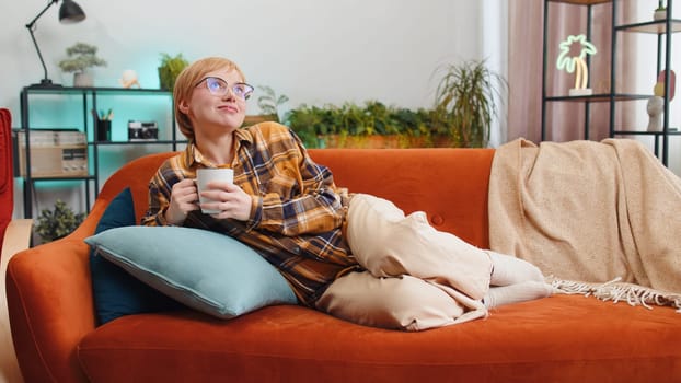 Portrait of attractive smiling young woman drinking a cup of warm coffee or herbal tea lying on sofa in the morning. Girl at home living room apartment enjoying comfortable relaxing resting alone