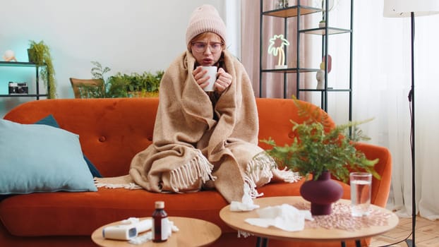 Sick young woman wear hat wrapped in plaid sit alone shivering from cold on sofa drinking hot tea in unheated apartment. Unhealthy ill girl in glasses feeling discomfort try to warming up at home room