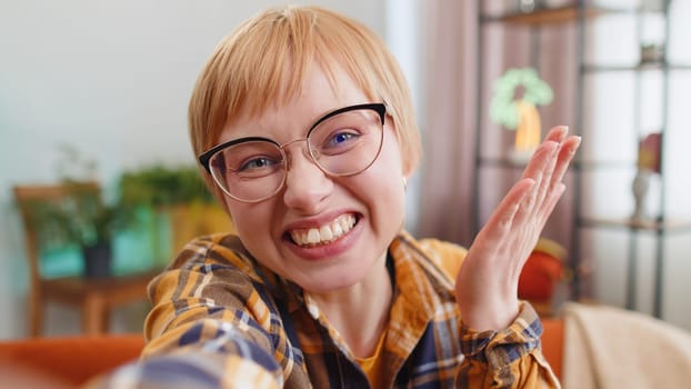 POV portrait of young woman blogger taking selfie on smartphone recording communicating video call online with subscribers sitting on sofa. Girl in glasses laughing waving hello at home room apartment
