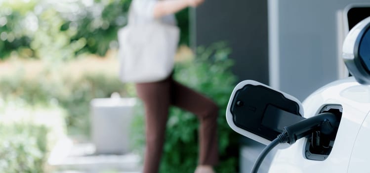 Focus EV charger plugged into EV car at home charging station with blurred background of progressive woman walking in background. Elective vehicle powered by clean energy for eco-friendly concept.