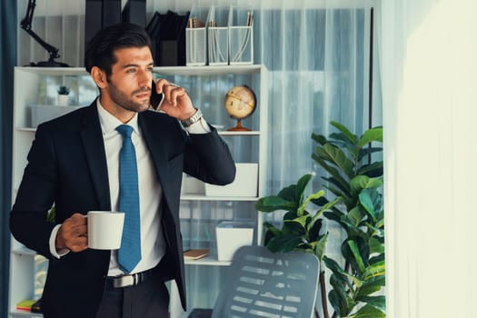 Diligent and busy businessman talking on phone call with coworker in modern office. Making sales call to client with coffee in hand, standing posing confidently as professional office worker. Fervent