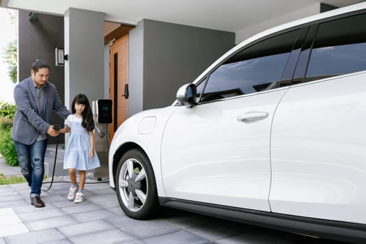 Progressive father and daughter plugs EV charger from home charging station to electric vehicle. Future eco-friendly car with EV cars powered by renewable source of clean energy.