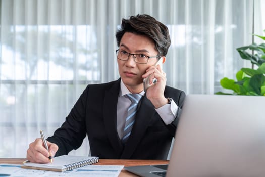 Businessman working on laptop for on office desk workspace with his smartphone. Smart executive researching financial business data or planning strategic business, talking on phone. Jubilant