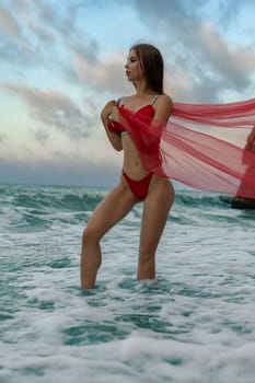 A sexy fitness model girl in a red swimsuit stands in the sea, wrapped in a transparent red cloth