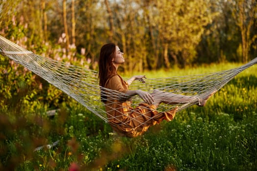 a happy woman in a long orange dress is resting sitting in a hammock at the dacha, smiling pleasantly looking away, illuminated by the summer sun during sunset. Horizontal photo. High quality photo