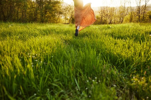 horizontal photo of a woman in an orange dress photographed without a face, standing in a green field during sunset, illuminated from the back. High quality photo