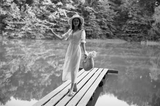 monochrome photo of a woman on the pier of a forest lake with a wicker hat looking at the camera. High quality photo
