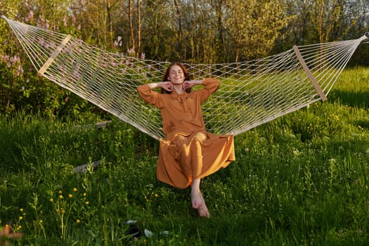 horizontal photo of a beautiful, red-haired woman lying in a hammock enjoying a rest in a long orange dress, on a warm summer day, touching her hair with her hand. High quality photo