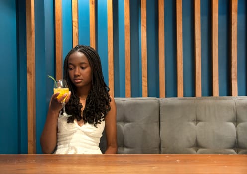 copyspace of afro descendant girl on a blue sofa looking with a puzzled look at her cocktail. High quality photo
