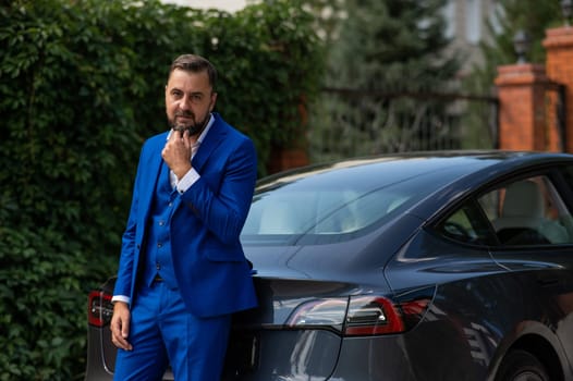 Caucasian bearded man in a blue suit stands near a black car in the countryside in summer