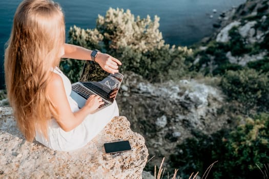 Successful business woman in yellow hat working on laptop by the sea. Pretty lady typing on computer at summer day outdoors. Freelance, travel and holidays concept.