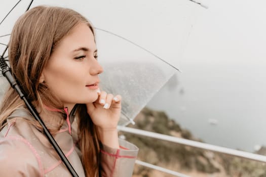 Woman rain park. Happy woman portrait wearing a raincoat with transparent umbrella outdoors on rainy day in park near sea. Girl on the nature on rainy overcast day