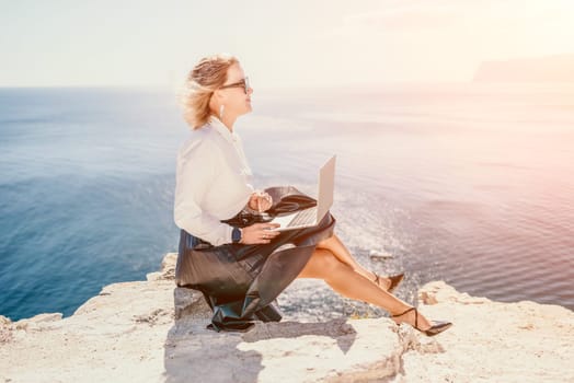 Digital nomad, Business woman working on laptop by the sea. Pretty lady typing on computer by the sea at sunset, makes a business transaction online from a distance. Freelance, remote work on vacation