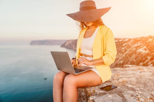 Successful business woman in yellow hat working on laptop by the sea. Pretty lady typing on computer at summer day outdoors. Freelance, travel and holidays concept.