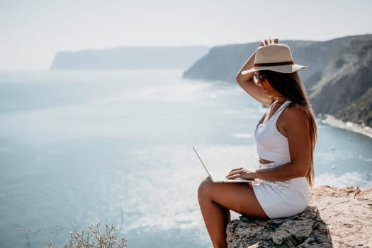 Successful business woman in yellow hat working on laptop by the sea. Pretty lady typing on computer at summer day outdoors. Freelance, travel and holidays concept.