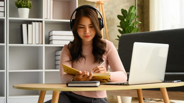 Pretty young asian woman wearing headphone sitting front of laptop and writing notes, planning workday in personal daily planner.