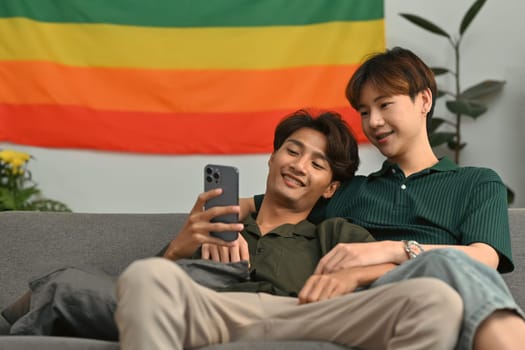 Beautiful moment of happy same sex male couple checking social media on smartphone while relaxing on couch at home.