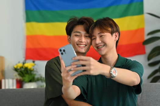 Joyful gay couple taking a selfie with smartphone in living room rainbow flag in background. LGBT, love and human rights concept.