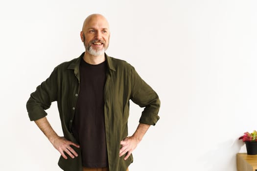 Smiling mid-aged bald man with silver beard standing confidently in front of white wall. His positive posture and expression radiate happiness and joy, showcasing contentment and cheerful demeanor. . High quality photo